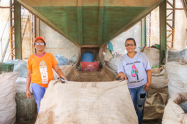 A foto mostra Miriam Silva Benvindo, cooperada da Cooprelp, e Catarina de Fátima Vaz, presidente da cooperativa de reciclagem, segurando um saco utilizado para a coleta de materiais recicláveis,