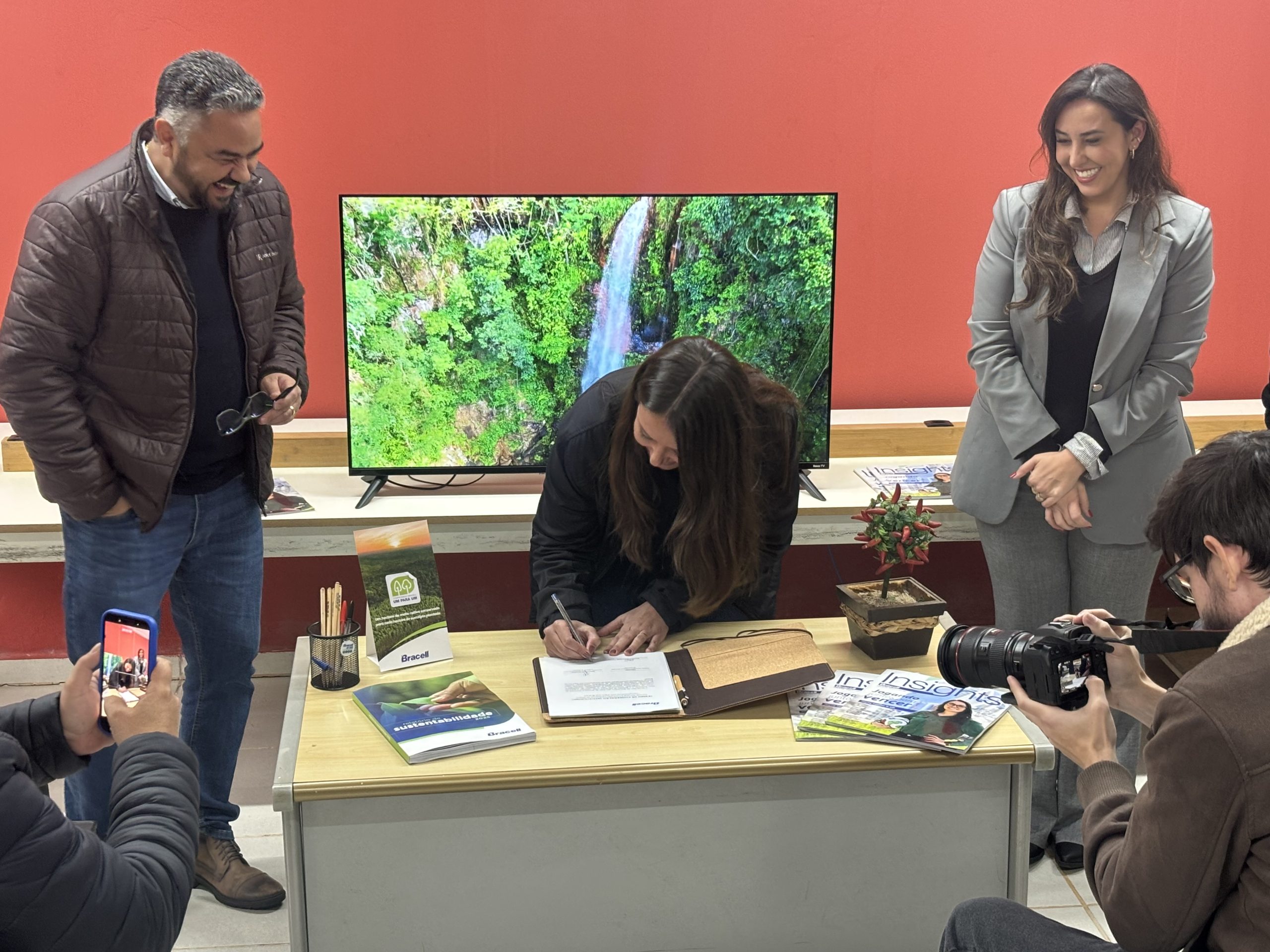 Pessoa assina documento sobre mesa durante evento de formalização de parceria entre Bracell e Prefeitura de Botucatu. Ao redor, outras pessoas acompanham o momento, enquanto um monitor ao fundo exibe imagem de uma cachoeira em área verde.