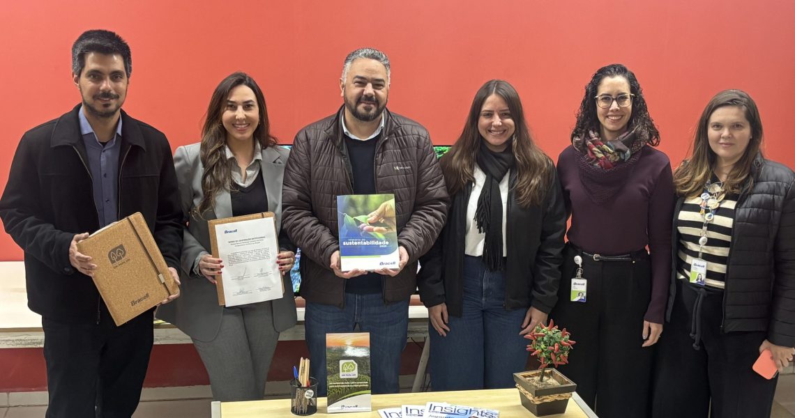 Seis pessoas posam lado a lado em ambiente interno durante oficialização de parceria entre a Bracell e a Prefeitura de Botucatu. Algumas seguram documentos e materiais institucionais relacionados ao Compromisso Um-Para-Um, em frente a uma mesa com revistas e itens de escritório.