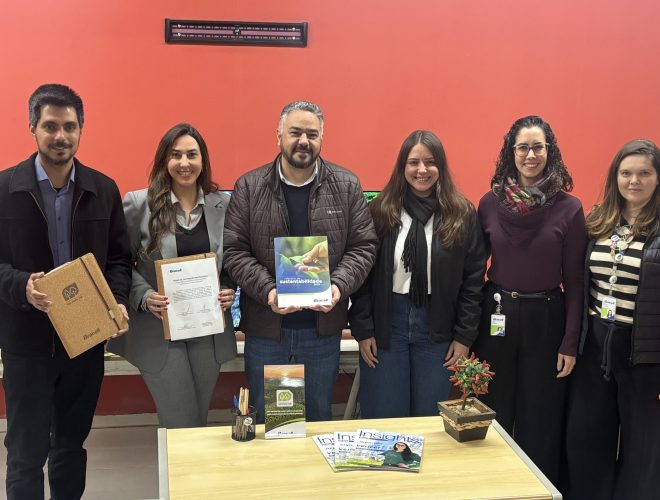 Seis pessoas posam lado a lado em ambiente interno durante oficialização de parceria entre a Bracell e a Prefeitura de Botucatu. Algumas seguram documentos e materiais institucionais relacionados ao Compromisso Um-Para-Um, em frente a uma mesa com revistas e itens de escritório.