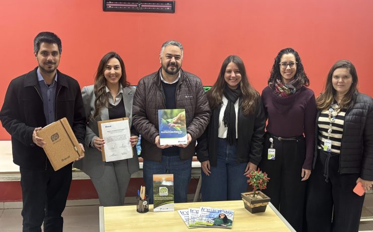 Seis pessoas posam lado a lado em ambiente interno durante oficialização de parceria entre a Bracell e a Prefeitura de Botucatu. Algumas seguram documentos e materiais institucionais relacionados ao Compromisso Um-Para-Um, em frente a uma mesa com revistas e itens de escritório.
