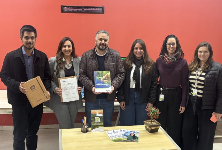 Seis pessoas posam lado a lado em ambiente interno durante oficialização de parceria entre a Bracell e a Prefeitura de Botucatu. Algumas seguram documentos e materiais institucionais relacionados ao Compromisso Um-Para-Um, em frente a uma mesa com revistas e itens de escritório.