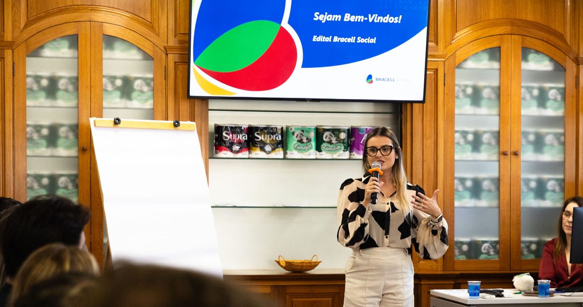 Pessoa faz apresentação durante evento do Edital Bracell Social em uma sala de reuniões. Ao fundo, uma tela exibe a mensagem “Sejam Bem-Vindos! Edital Bracell Social”, enquanto participantes acompanham a atividade.