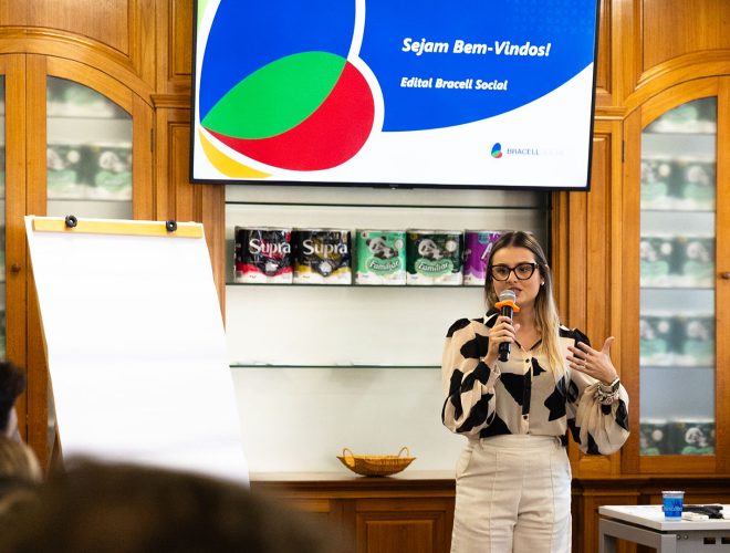 Pessoa faz apresentação durante evento do Edital Bracell Social em uma sala de reuniões. Ao fundo, uma tela exibe a mensagem “Sejam Bem-Vindos! Edital Bracell Social”, enquanto participantes acompanham a atividade.