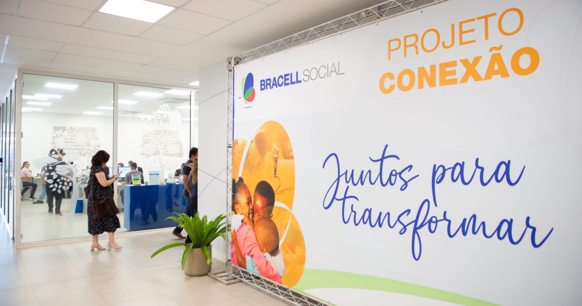 Painel do Projeto Conexão da Bracell Social em espaço de eventos, com participantes ao fundo durante atividade voltada ao fortalecimento de organizações sociais.