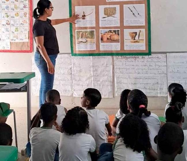 Educadora apresenta painel com atividades em sala de aula para estudantes sentados no chão.