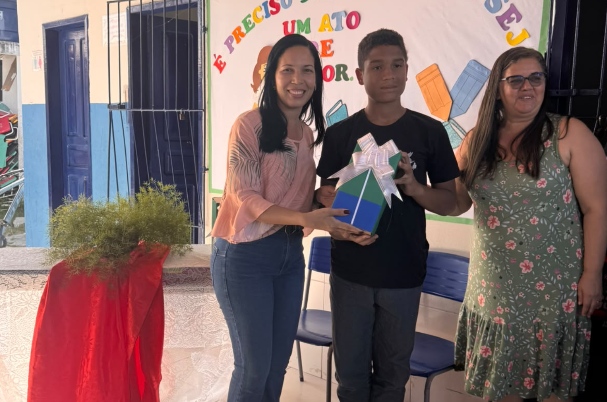 Estudante recebe premiação durante cerimônia escolar acompanhado por duas pessoas em espaço educativo.