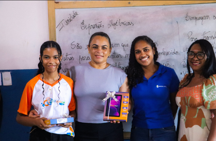 Estudante e três participantes exibem premiação em sala de aula diante de quadro com conteúdo escolar.